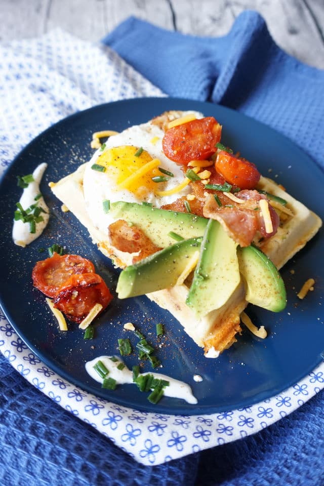 herzhafte Frühstückswaffeln auf blauem Teller, belegt mit Avocado, gebratenen Tomaten, einem Spiegelei, geriebenem Käse und gebratenem Baconscheiben - BLW Rezepte