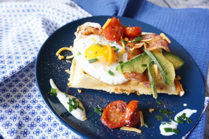 blauer Teller mit einem Waffel, die mit Spiegelei, Tomaten, Avocado und Frühstücksspeck ganiert ist. Außerdem Frische Kräuter und Creme fraiche. Frühstückswaffel als Frühstück für Babys, Kleinkinder un ddie ganze Familie