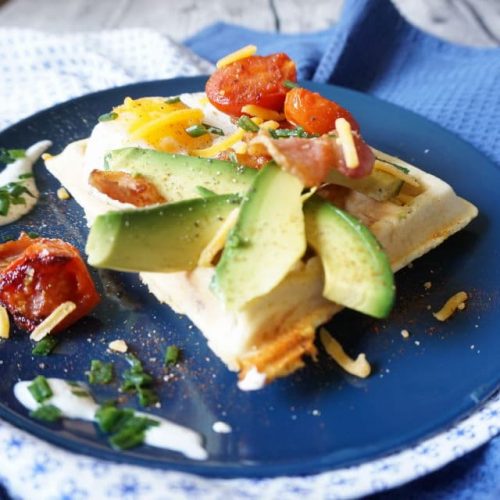blauer Teller mit rechteckiger herzhafter Frühstückswaffel ganiert mit Avocadoschnitzen, Coktailtomaten, Spiegelei, geriebenem Käse und Bacon