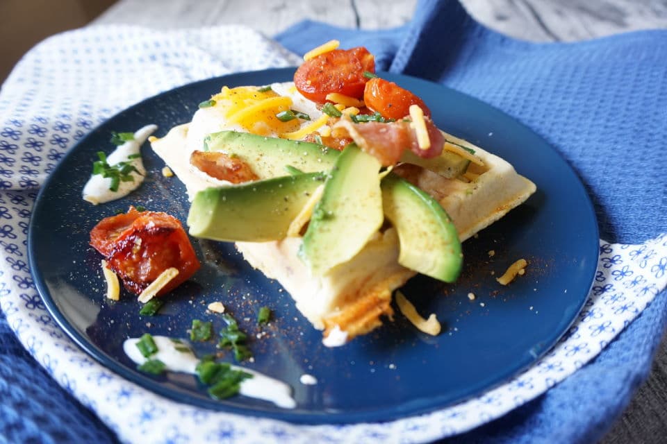 blauer Teller mit rechteckiger herzhafter Frühstückswaffel ganiert mit Avocadoschnitzen, Coktailtomaten, Spiegelei, geriebenem Käse und Bacon