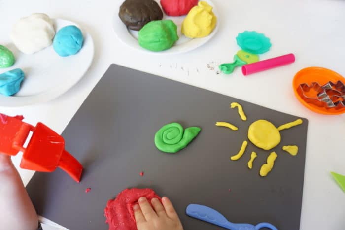 graues Brett mit einer Sonne aus gelber Knete und einer grünen Schnecle, verschiedene selbstgemachte Knete auf einem Teller