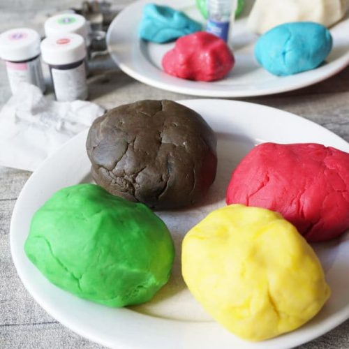 Knete selber machen one Alaun - zwei weiße Teller mit Knete in verschiedenen Farben - gelb, pink, grün, braun und grün auf einem Teller, Gllitzerknete auf dem anderen Teller - nebendran Lebensmittelfarben