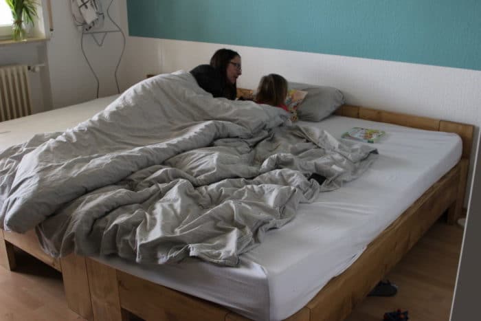 Mama und Tochter liegen auf dem Bauch im großen Familienbett unter der Decke - graue Bettwäsche - lesen zusammen ein Buch zum Thema Schwangershcaft und Geschwister