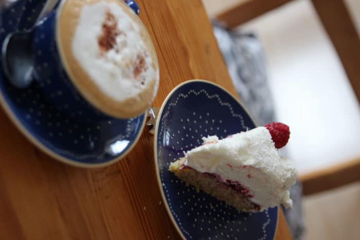 Milchkaffee mit schönem Schaum in einer blauebn Kaffeetasse auf Unterteller nebem einem blauen Kuchenteller mit einem Stück Torte - Raffaellotorte mit Himbeeren - köstliche Kuchen und Torten fürs Wochenende, Geburtstag, Muttertag oder Valentinstag