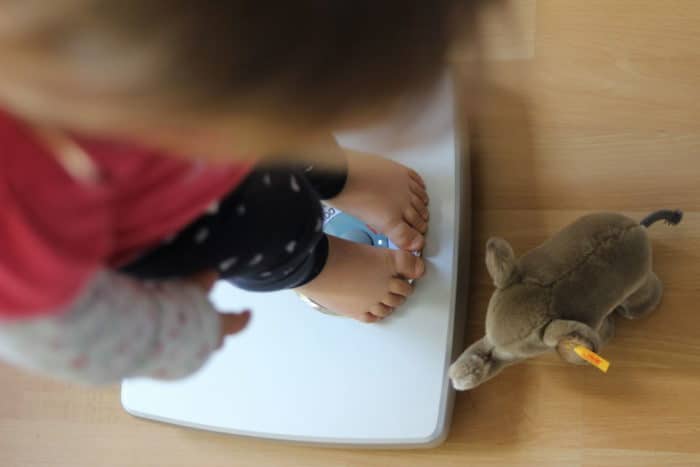 Mädchen steht auf der Waage mit kleinem Kuscheltier Elefant zu ihren Füßen - Wachstum bei Kindern im Blick behalten